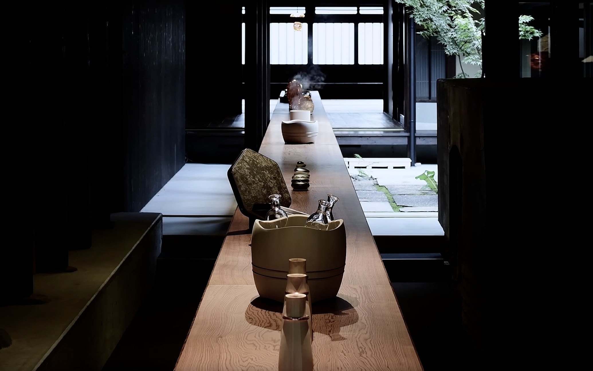 A narrow wooden counter with bottles in a ceramic holder, inside a dimly lit room leading to a brighter outdoor area with trees and a wooden deck.