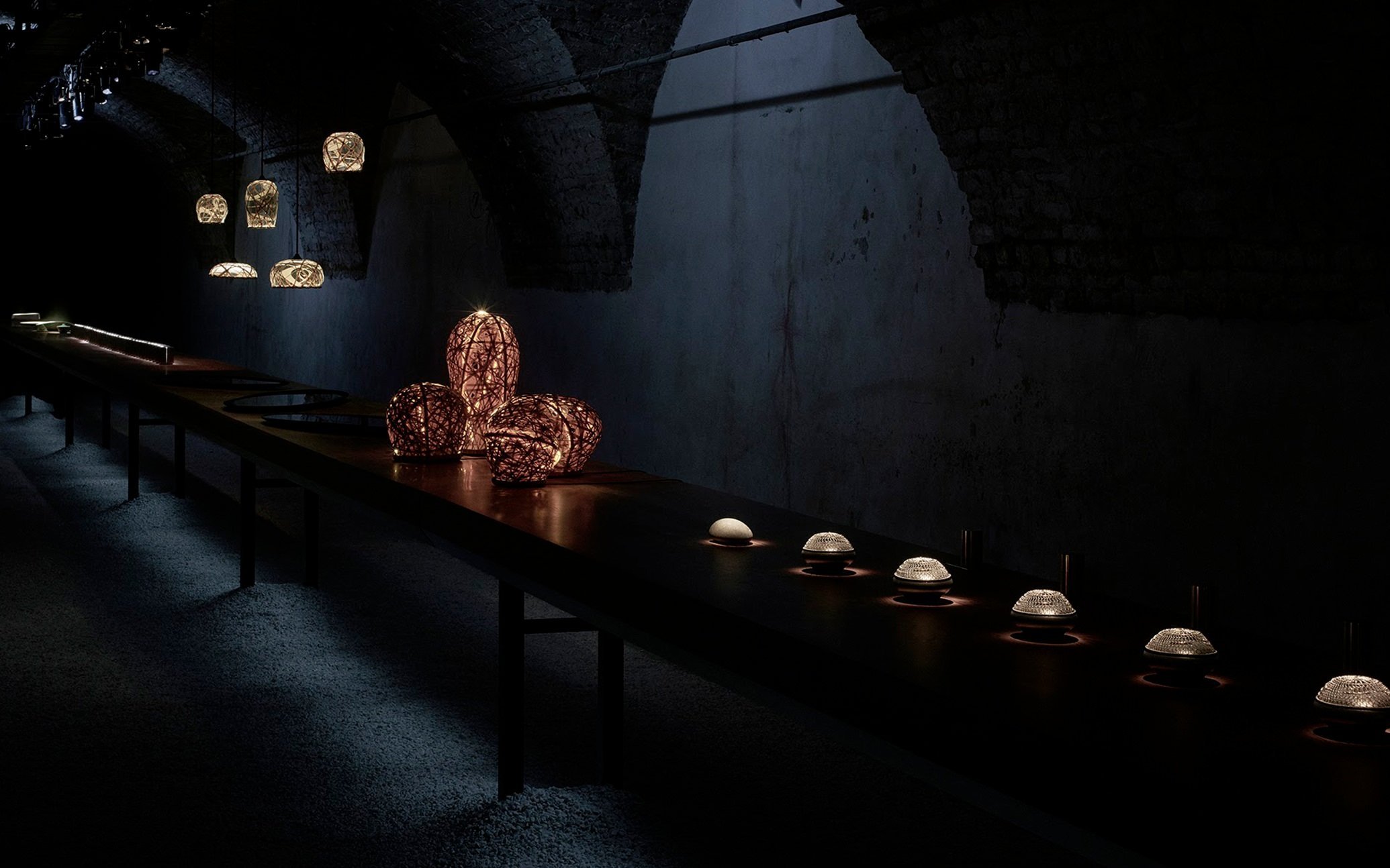 Dimly lit display of various decorative lamps and objects on a long wooden table inside a dark room.