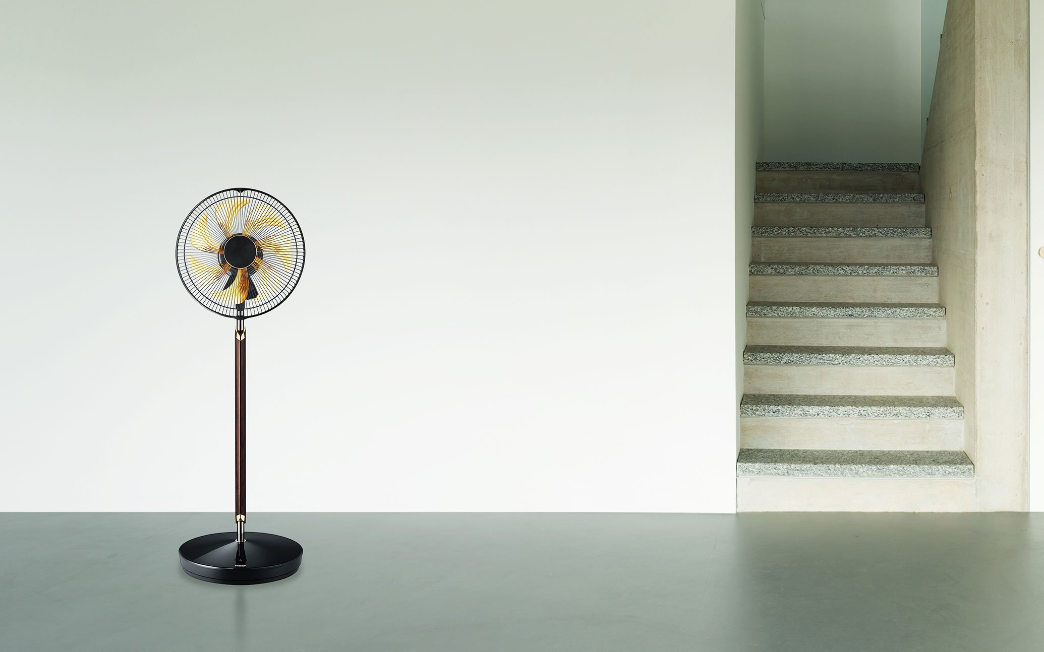 A black standing fan with yellow blades on a grey floor, next to a staircase with concrete steps and a white wall.