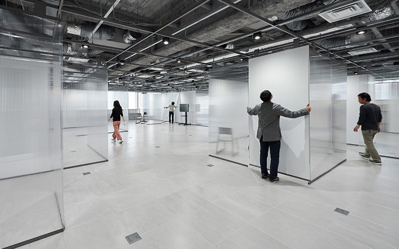 Spacious interior with transparent partitioned areas, exposed ceiling pipes and lighting, several people walking within the space