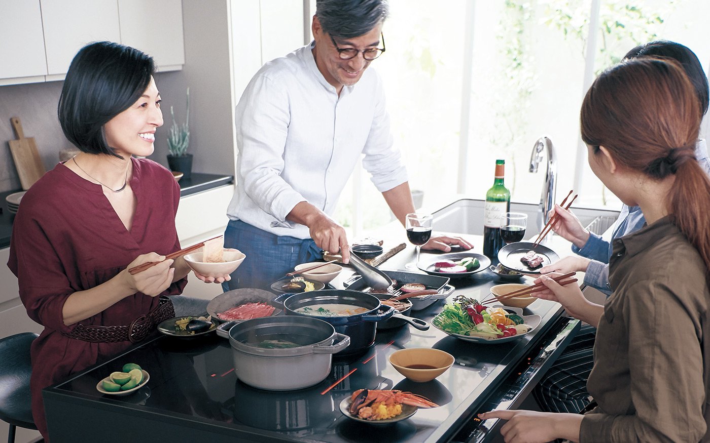 Dining scene with four people around a kitchen table. Pots and pans with cooking food, hands holding chopsticks, glasses of red wine, and plates with colorful ingredients.