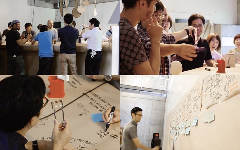 Group of men and women standing at a bar counter. Meeting scene with people writing notes and drawings on paper, and a workspace with sticky notes and memos on the wall.