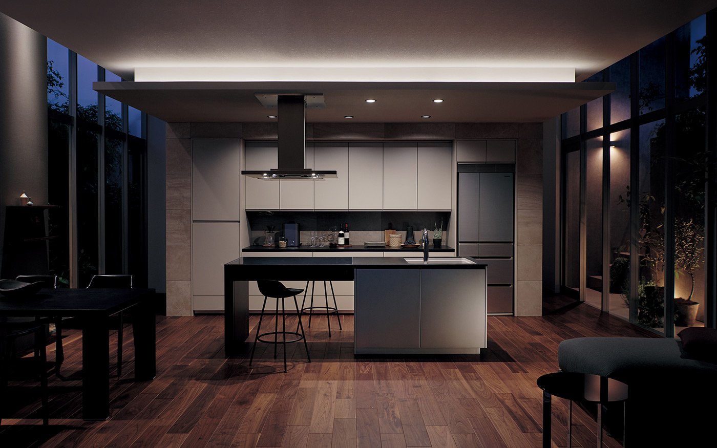 Modern gray kitchen surrounded by wood flooring. Central island counter with high chairs, cabinets and refrigerator in the back. Large glass windows and warm lighting.