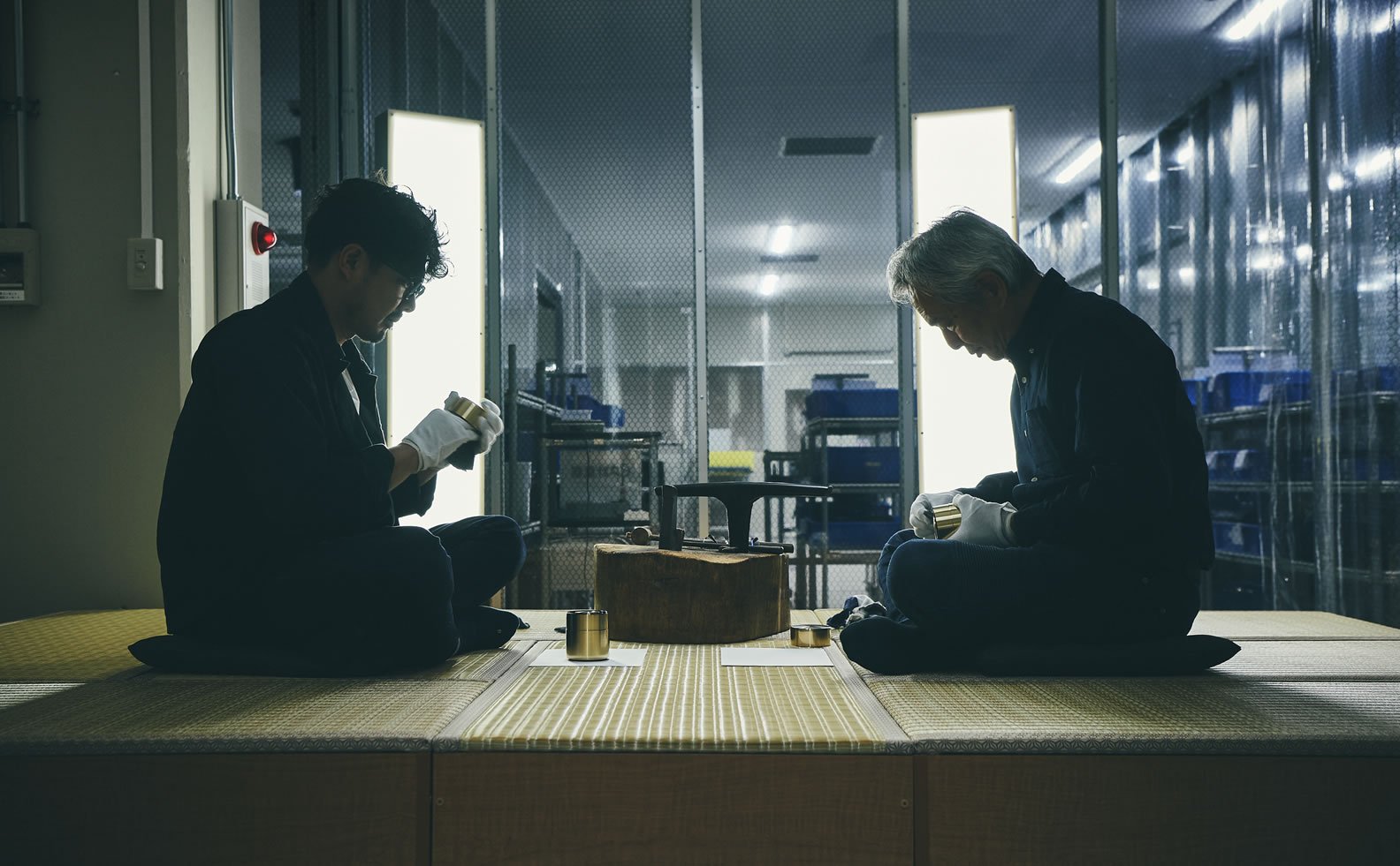 Two men sitting on a tatami mat with a cylindrical speaker between them, factory partitions and blue storage shelves in the background
