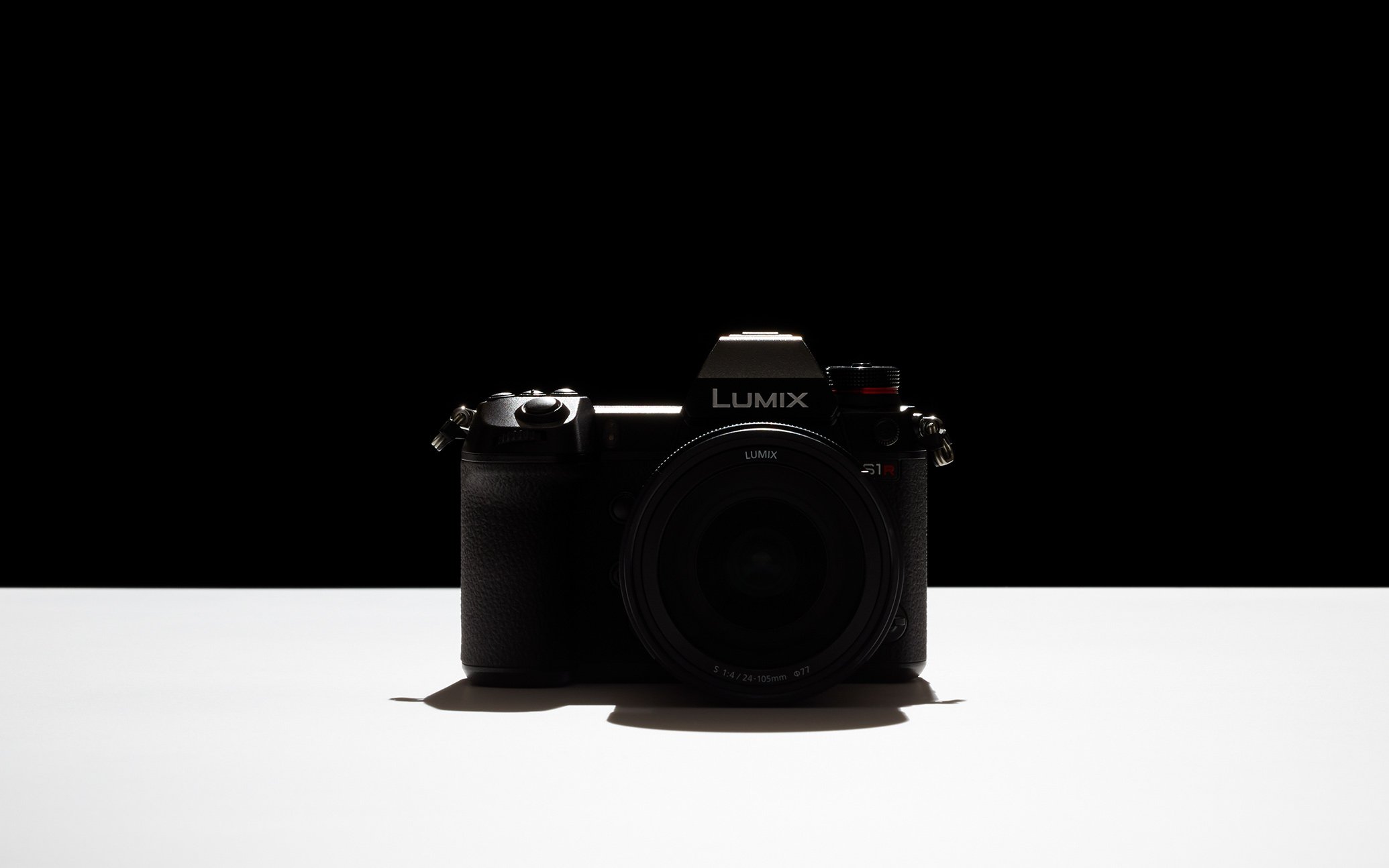 Black Panasonic Lumix camera body centered with lens, placed on white surface, black background