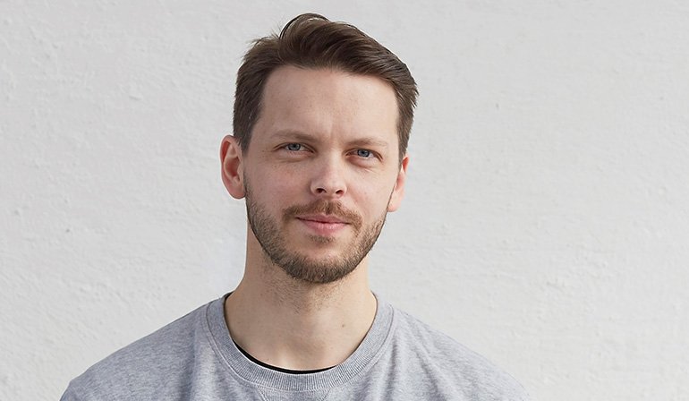 Upper body shot of a person wearing grey t-shirt. Short brown hair and beard. White background