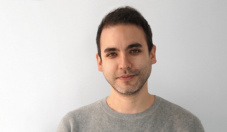 Upper body portrait against white background. Person wearing grey knit sweater. Short dark hair and beard.
