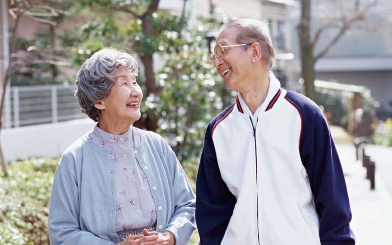 Walking path with greenery outdoors. Two elderly people in athletic wear. One wearing white and navy jacket, another in light blue cardigan with floral top.
