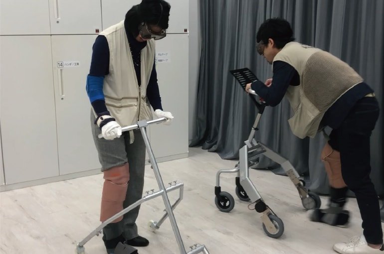 Indoor walking aid test scene. Two people wearing beige vests using walking aids. Silver walking aids in room with white walls and grey curtains. Checking operation on tablet device