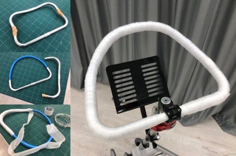 Walking aid handlebar prototype. Metal frame covered with white cushioning material. Black tablet holder attached. Parts on green cutting mat and assembled final product