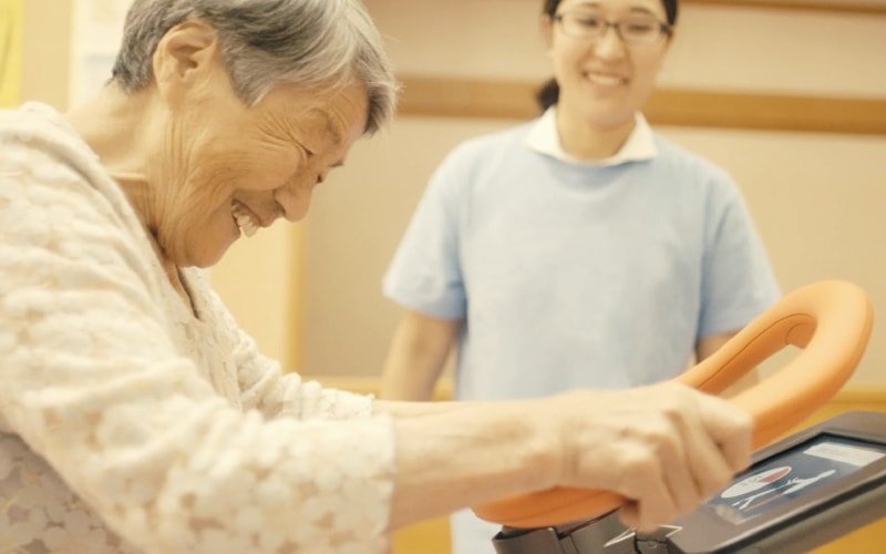 Indoor scene with walking aid operation. Elderly person in white floral clothing using walking aid with orange handlebar and tablet screen. Staff member in light blue uniform in background