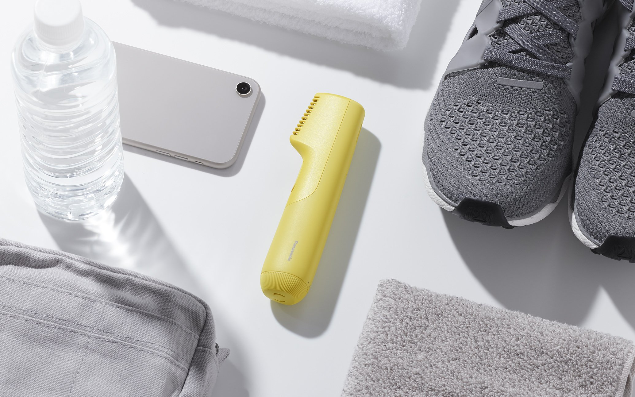 Photo showing a gray smartphone, yellow trimmer, clear water bottle, white towel and bath towel, gray sneakers on a white surface