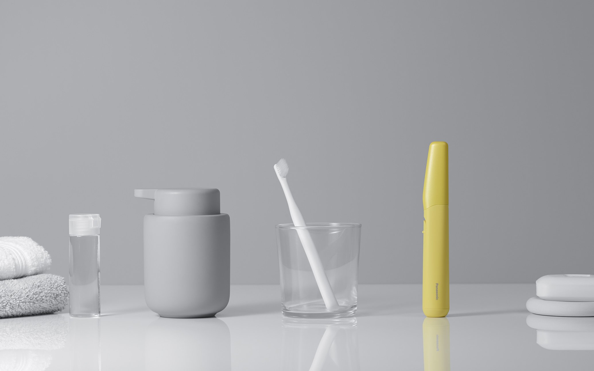 Image of a gray container, white toothbrush, yellow electric toothbrush, white towels, and water bottle on a bathroom counter