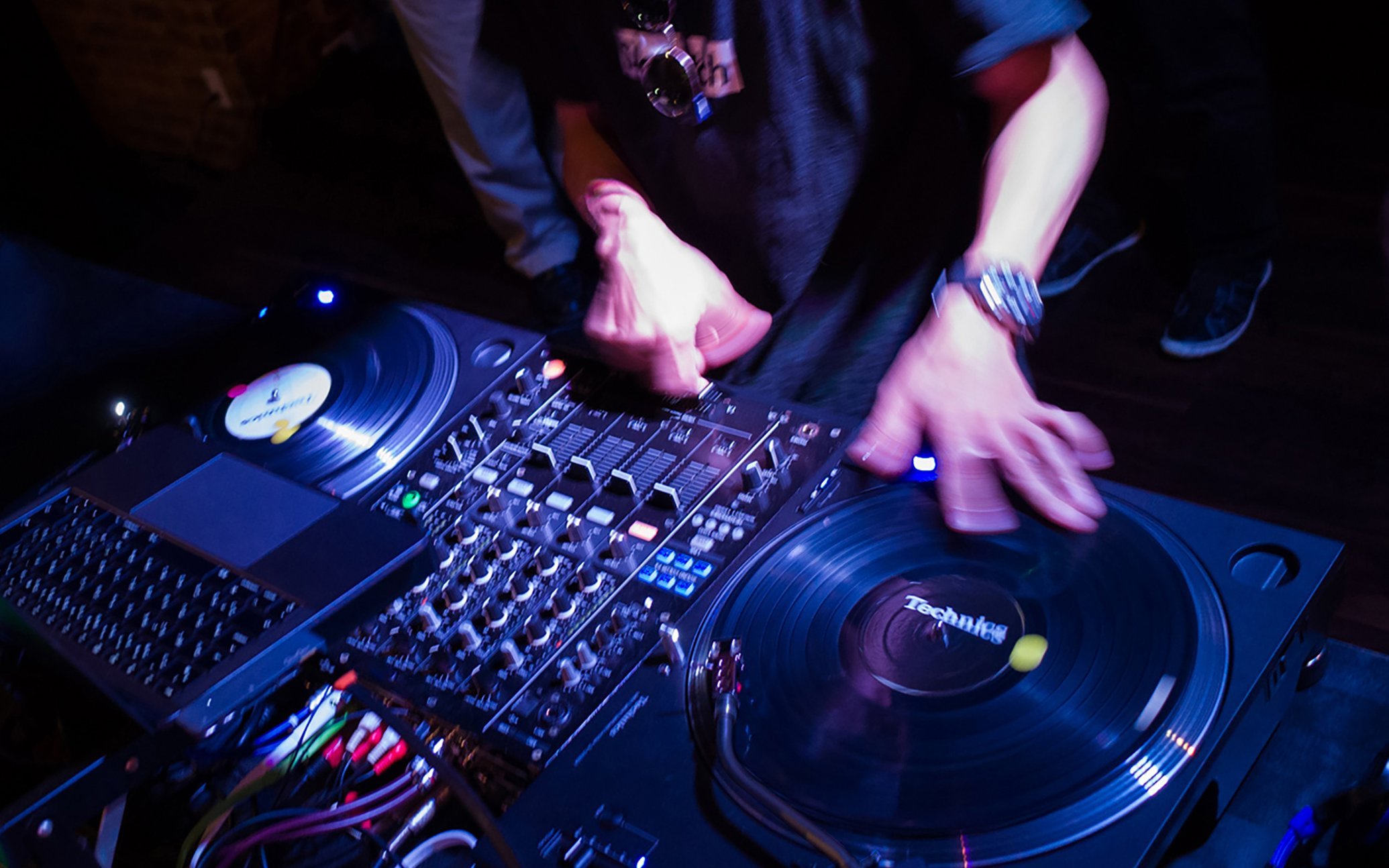Turntables and mixer with hands operating, DJ equipment on a dark surface.