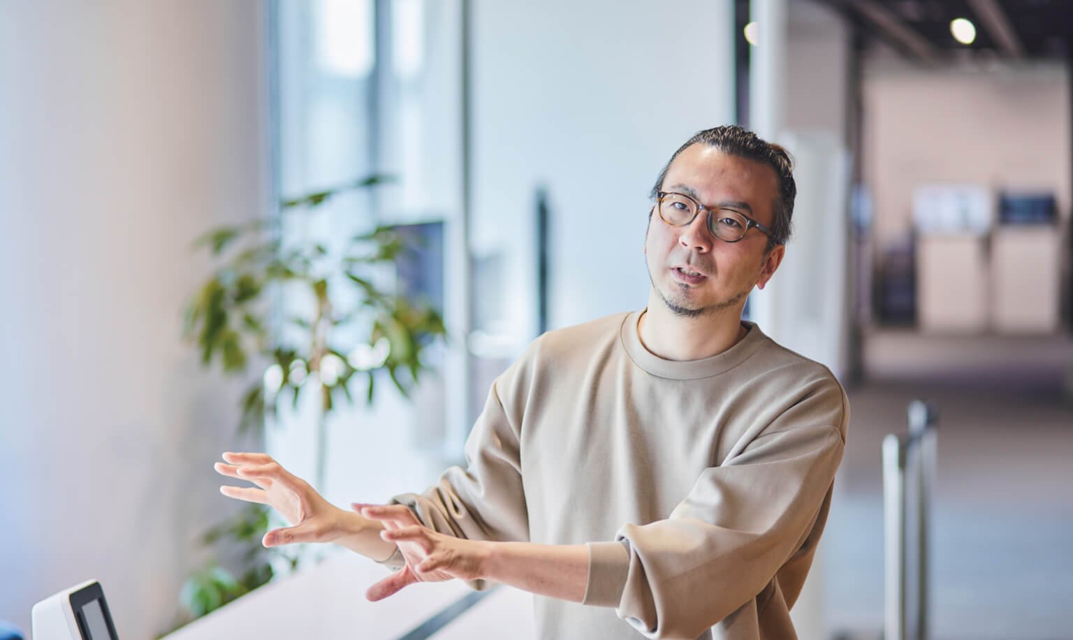 A person with blurred face gesturing with both hands in a modern indoor setting near a window.