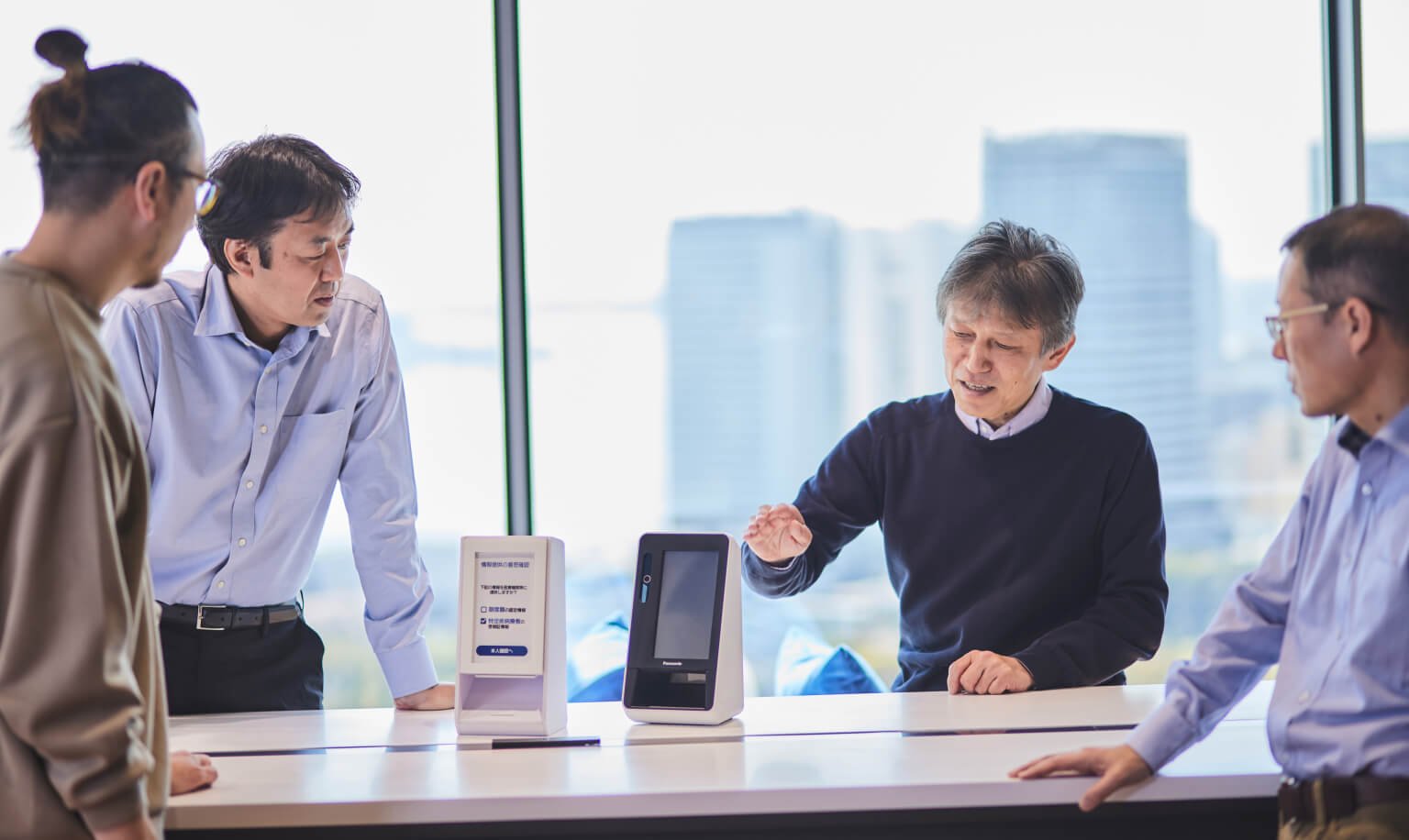 Four businesspeople gathered around a table in a conference room with large windows, a card reader and a paper model placed on the table, high-rise buildings and blue sky visible outside.