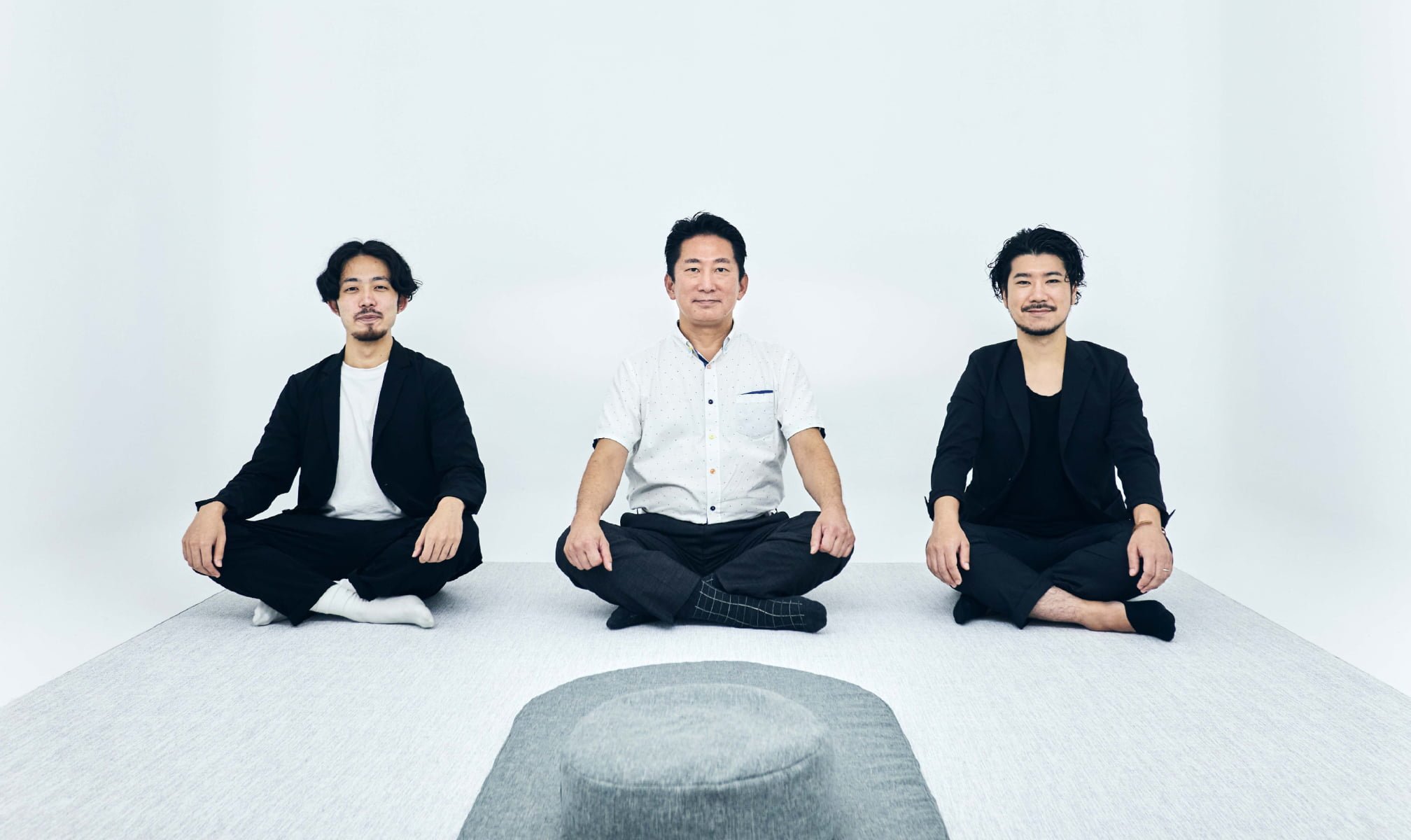 Three people sitting on the floor against a white background, the person in the middle is male, flanked by females, faces blurred