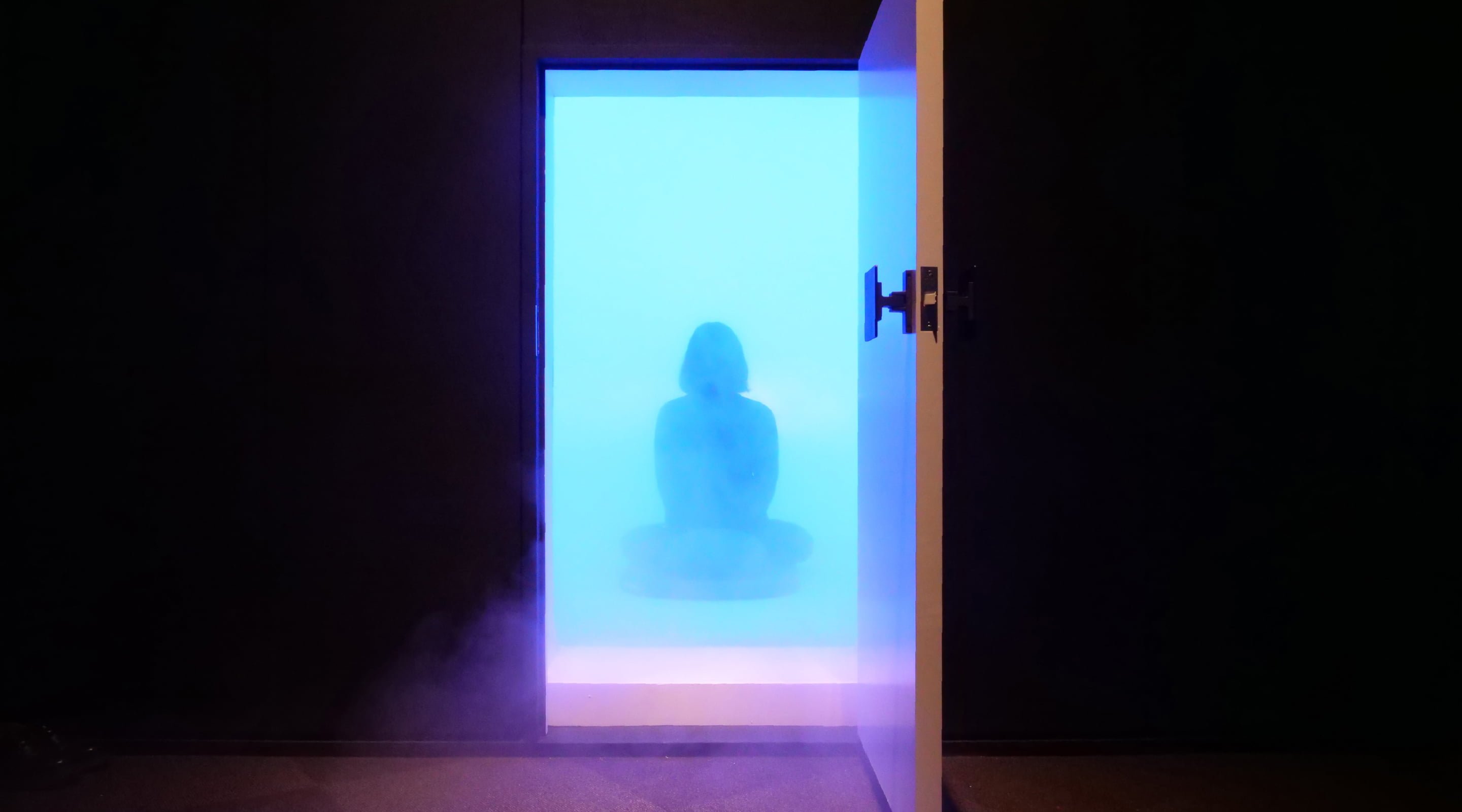 Figures sitting in a space surrounded by blue light, open door, foggy effect, dark background.