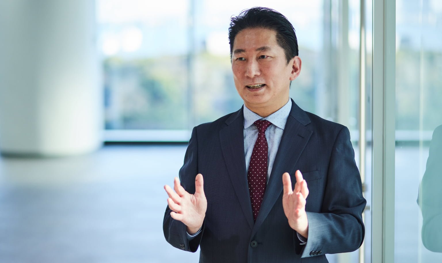 Businessman wearing a dark suit, white shirt, red polka dot tie, standing near window inside, hands raised as if explaining.