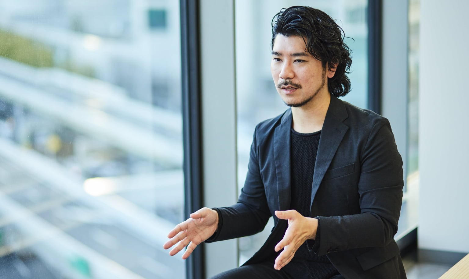 Man sitting by the window inside, wearing a black suit and black sweater, gesturing while talking