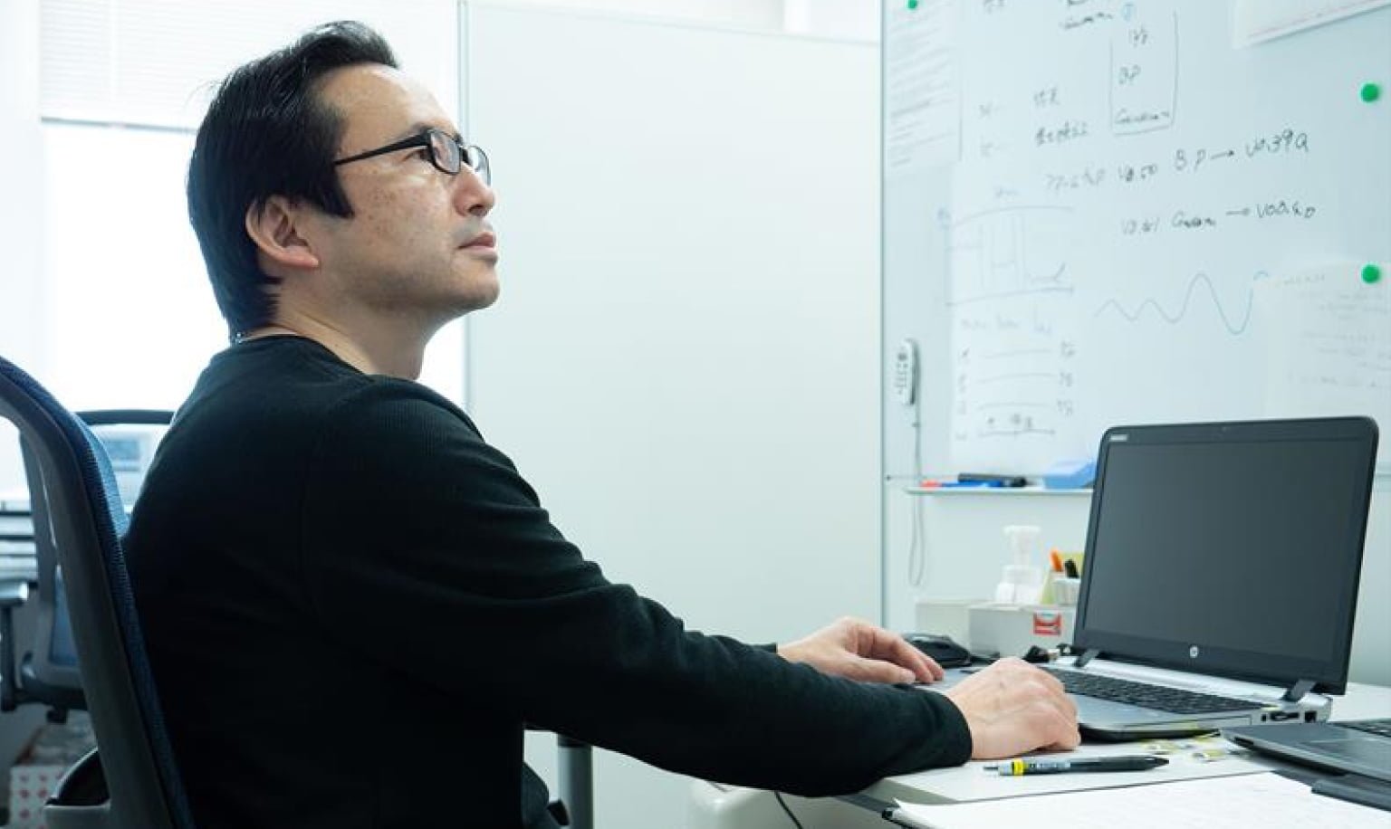 Man working at white desk, wearing a black long-sleeve shirt, with a laptop and stationery