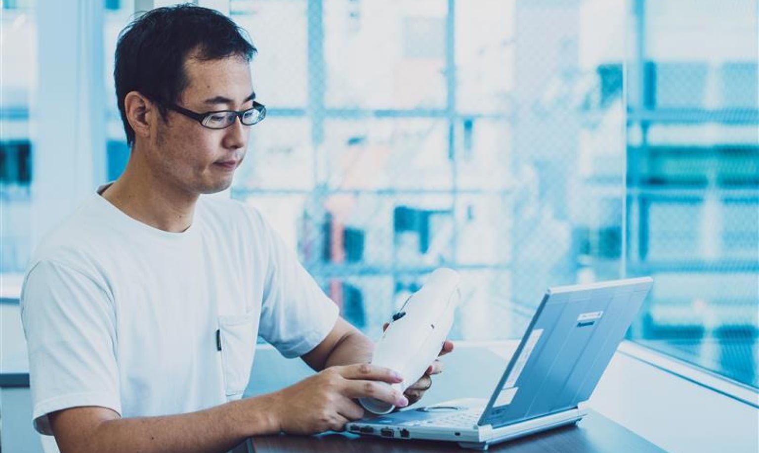 Man in a white shirt holding documents in hand, working on a laptop by the office window