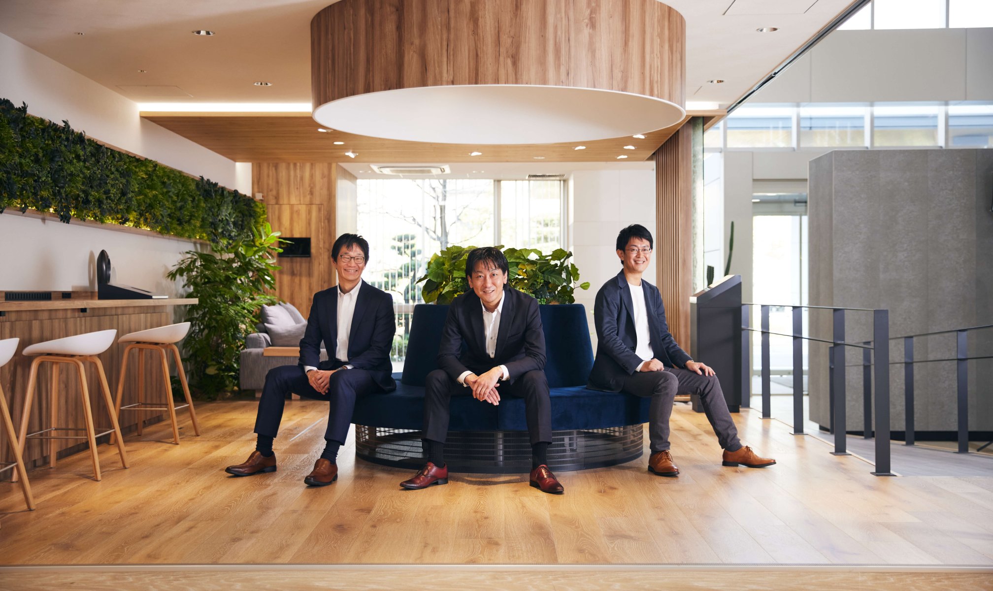 A space with a central blue circular sofa topped by a large hood, where three businessmen are seated. The background features green indoor plants, a large window, and natural sunlight streaming in.