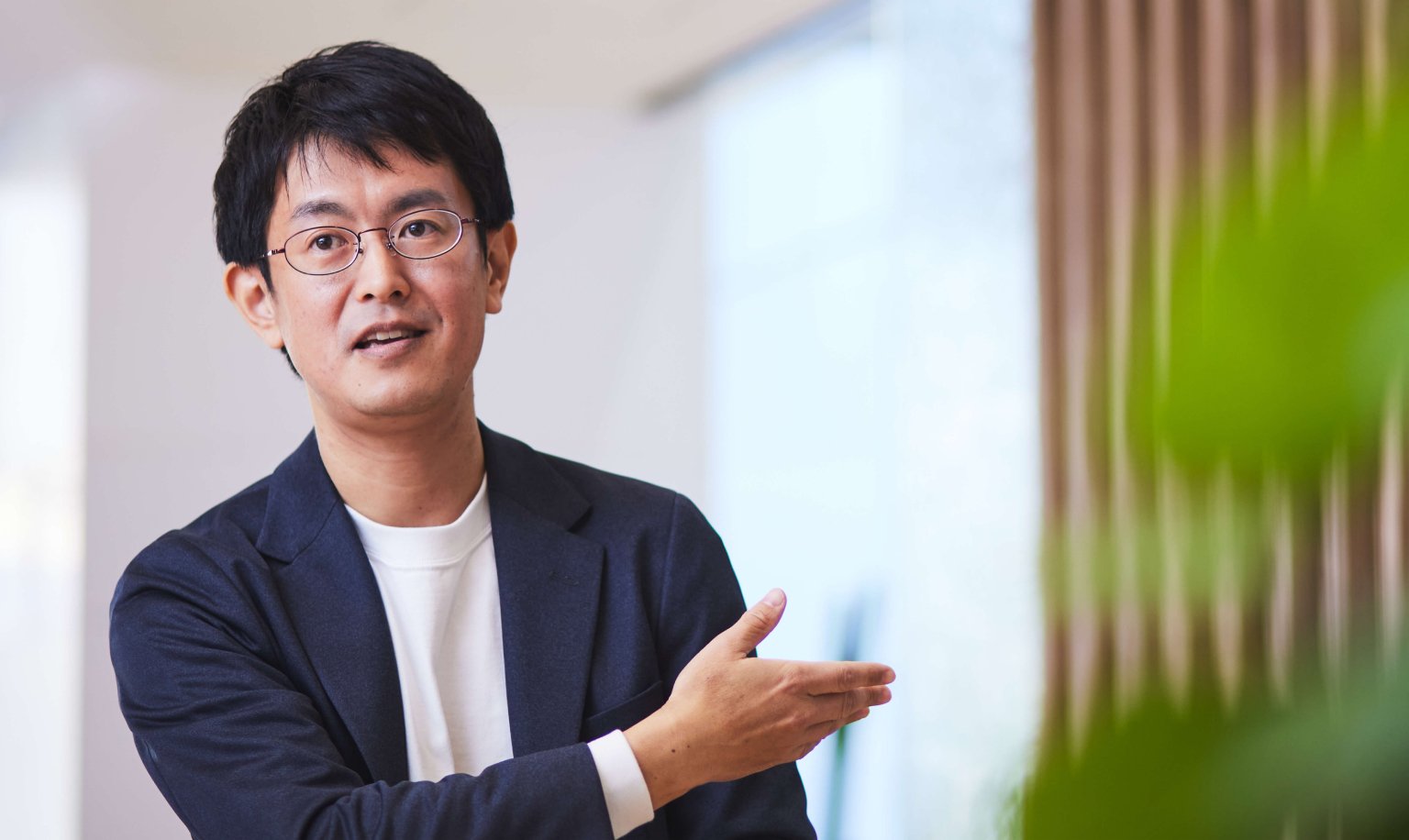 A man wearing glasses, a T-shirt, and a jacket is speaking while extending his right hand.