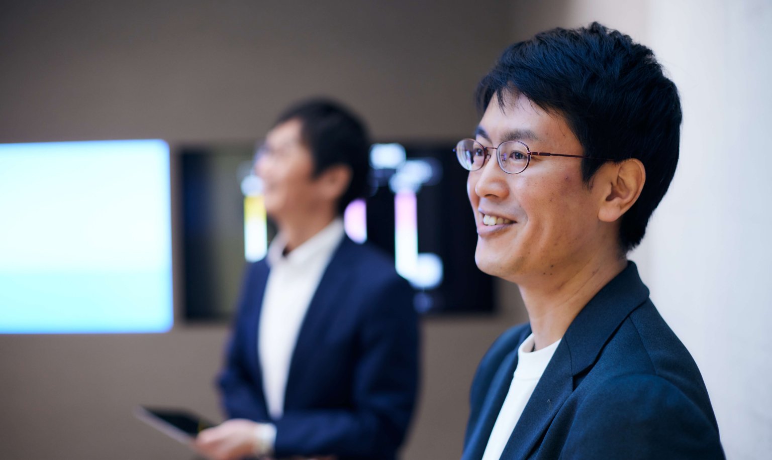 Two businessmen wearing white shirts and navy jackets are seated in a conference room. The background shows a screen and a wall.