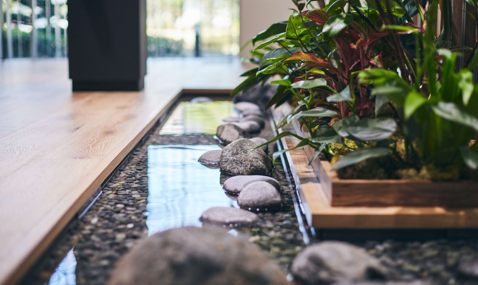 A small pond with flowing water and stones arranged along a wooden floor, accompanied by a wooden planter filled with green indoor plants beside it.