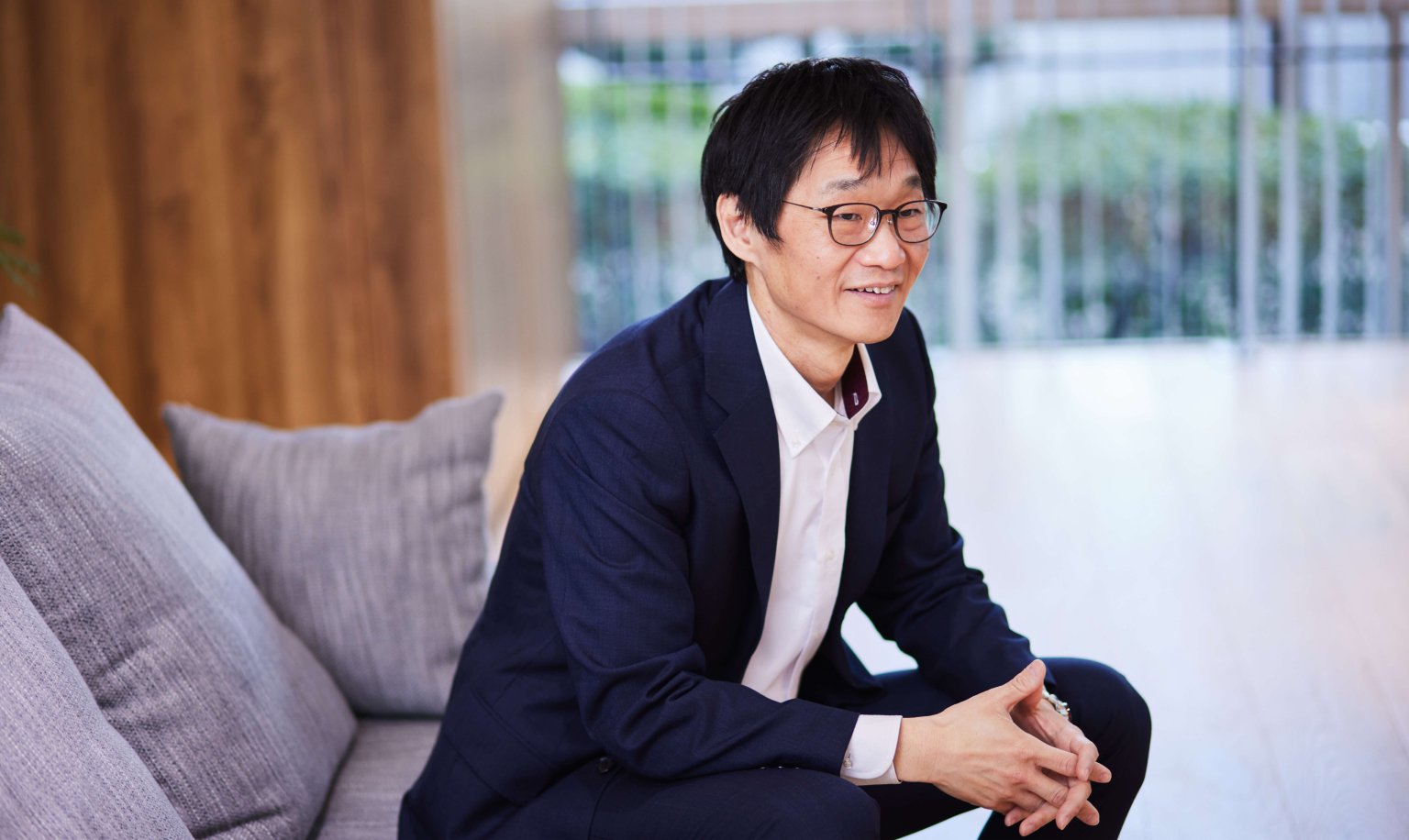 A man wearing glasses, a white shirt, and a navy blazer is sitting on a sofa with his hands clasped together.
