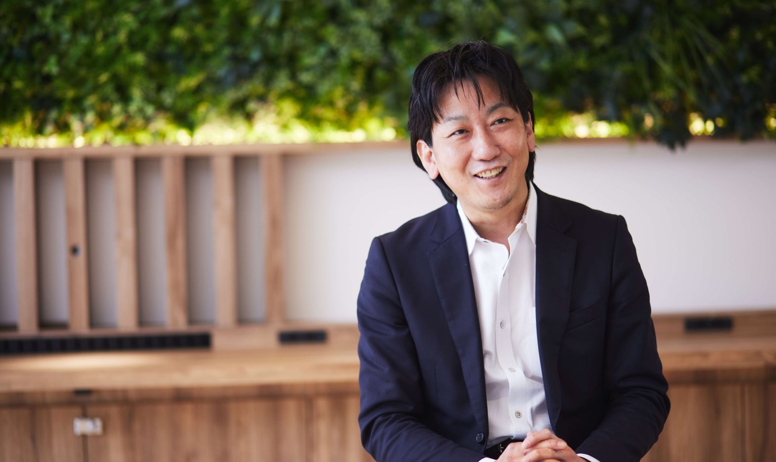 A man wearing a white shirt and navy blazer is sitting against a background of wooden panels and greenery, with his hands clasped together.
