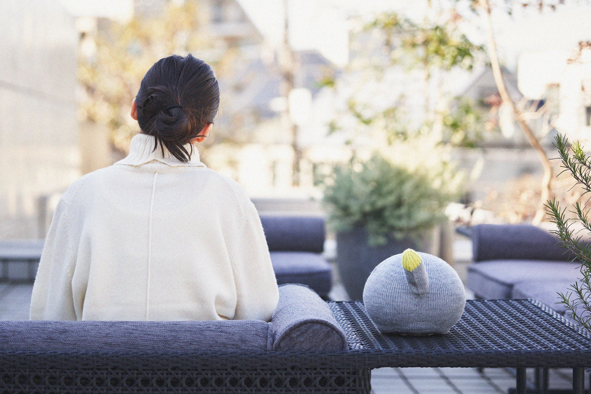 Woman sitting on outdoor sofa on balcony, with a gray knitted plush hat and plants in background