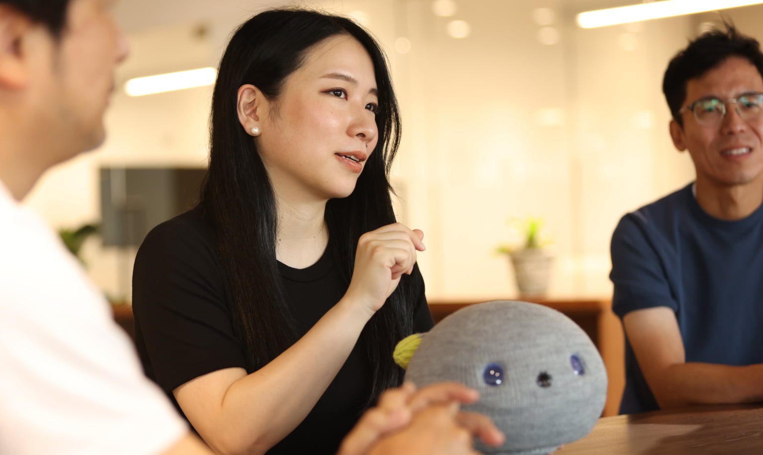 A woman with long black hair is sitting in front of NICOBO, which is placed on the table, and talking.