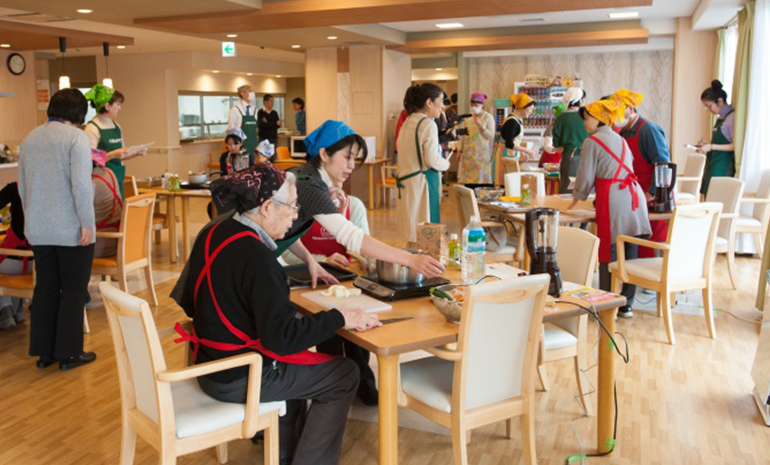 Photo : At a serviced residence for the elderly, Yoko Nakao chats with residents in a cooking class using electronic appliances. 